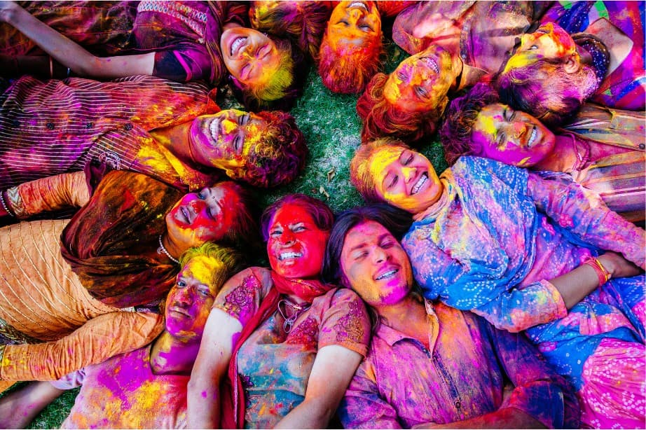 Friends lying in a circle on the grass, faces and clothes covered in bright Holi powder