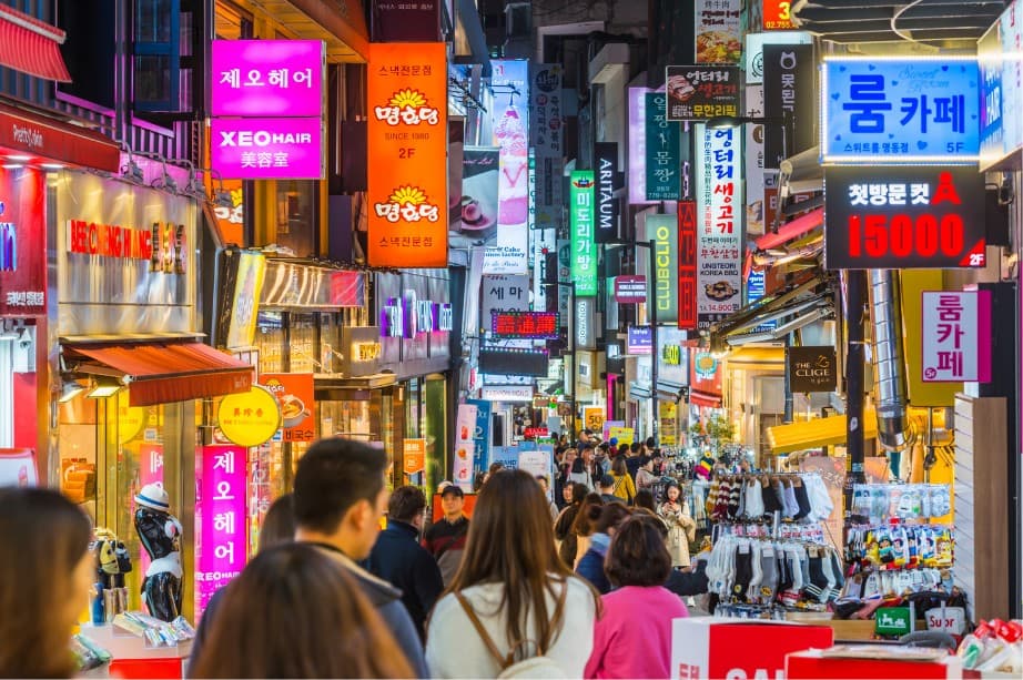 A crowded shopping street in Myeongdong, Seoul, lined with bright neon signs at dusk